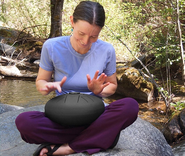 a woman playing a steel tongue drum by a flowing creek of water